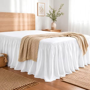 May include: A white bed valance with a gathered ruffle design draped over a bed frame. A tan knitted throw blanket is placed across the white bed sheets. The bed features a light wood headboard and is positioned in a room with light walls.