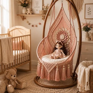 May include: A hanging macrame chair in dusty pink featuring a large central flower motif, a round white seat cushion, and long decorative fringe with wooden beads. A doll with brown hair wearing a pink dress sits in the center of the chair.