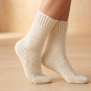 May include: A pair of cream-coloured, handmade crochet socks. The socks have a ribbed cuff and a textured stitch pattern throughout the body. The socks are displayed on a wooden surface.