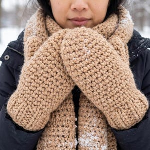 May include: A close-up of a tan crochet scarf and mittens. The mittens are thick and textured, with a ribbed cuff. The scarf is wrapped around the neck, and the background is a snowy outdoor scene. The mittens and scarf appear to be handmade.