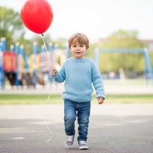 以下が含まれることがあります： 水色のセーターとジーンズを着た幼い子供が赤い風船を持っています。子供は公園の舗装された表面を歩いており、背景には遊び場が見えます。