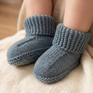 Op de afbeelding: Een paar babyslofjes in een zacht, gedempt blauw. De gebreide slofjes hebben een geribbelde manchet en een ronde neus. Ze rusten op een crèmekleurige gebreide deken. De slofjes lijken handgemaakt.