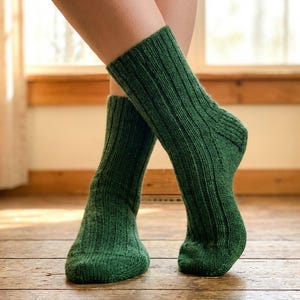 May include: A pair of green ribbed knit socks. The socks are a solid, deep green colour and appear to be made of a soft, warm material. The socks are worn on a person's feet, with the legs visible from the ankles down.