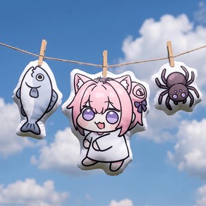 May include: Three plush toys hanging from a string against a blue sky. The toys include a cartoon fish, a character with pink hair and cat ears, and a spider. Each toy is attached to the string with a wooden clothespin.