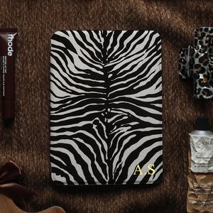May include: A collection of items including tortoise shell sunglasses, a book, a small glass bottle with coffee beans and a thank you tag, a Rhode lip tube, a zebra print e-reader case with gold A.S initials, a leopard print hair clip, a tube of cream, and a watch.