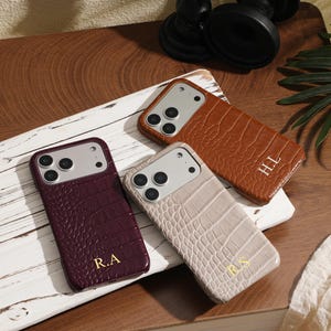 Op de afbeelding: Drie rechthoekige smartphonehoesjes met een gestructureerd krokodillenleerpatroon. Eén hoesje is donkerpaars met gouden initialen R.A, één is lichtbeige met de initialen R.S en één is lichtbruin met de gouden initialen H.L.