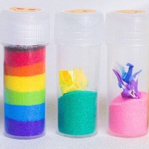 May include: Five clear plastic tubes with white lids, each containing different decorative elements. The tubes hold blue sand with shells, rainbow-layered sand, green sand with yellow paper, pink sand with a purple origami crane, and yellow glitter.