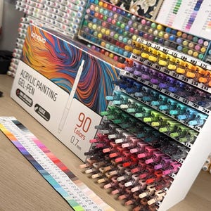 May include: A set of 90 acrylic painting gel pens with 0.7mm tips, organised in a tiered white display stand. The packaging features colourful abstract swirls and the text "Languo Acrylic Painting Gel-Pen 90 Colors 0.7mm". A colour swatch chart is included.