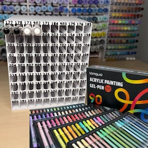Op de afbeelding: Een verzameling kunstbenodigdheden, waaronder gelpennen en stiften, georganiseerd in witte opbergrekken. De afbeelding toont een verscheidenheid aan kleuren en de tekst "Acrylic Painting Gel-Pen Plus 90".