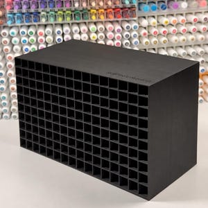 May include: A black rectangular marker organiser featuring a grid of square storage slots. The top surface is engraved with the text ArtProColoring. The unit is constructed with a matte finish and provides multiple individual compartments for pens.