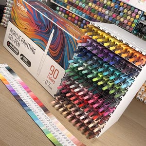 May include: A box of Languo acrylic painting gel pens with 90 colours, 0.7mm tips. The box features colourful abstract art. A colour chart is displayed next to the pens, showing the range of colours available.