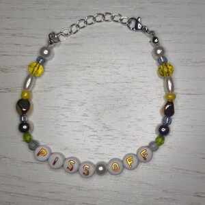 Puede incluir: Pulsera de cuentas con las palabras "PISS OFF" escritas en letras doradas sobre cuentas blancas. La pulsera presenta una mezcla de cuentas amarillas, grises y color perla, con una cadena y cierre plateados.