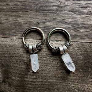 May include: A pair of silver-toned hoop earrings featuring decorative metal beads and a dangling clear, faceted crystal point pendant. Each hoop is circular with a hinged closure, accented by two filigree-style beads and a smooth central bead.