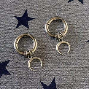 May include: A pair of silver-toned hoop earrings featuring a hinged closure mechanism. Each hoop is adorned with a small, textured metal ring and a dangling crescent moon charm. The metal has a polished, reflective finish throughout the design.