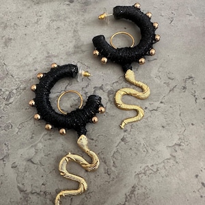Può includere: Un paio di orecchini a cerchio neri con ciondoli a forma di serpente dorati. I cerchi sono avvolti in filo nero e adornati con piccole perle dorate. Gli orecchini sono esposti su una superficie grigia.