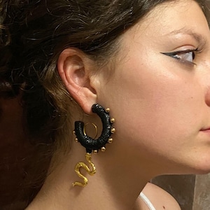 May include: Black hoop earrings with gold detailing and a gold snake pendant. The earrings feature a wrapped design with small gold studs. The snake charm hangs from the bottom of the hoop.