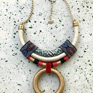May include: A statement necklace featuring a gold-tone chain and a large circular pendant wrapped in gold thread. The collar section includes segments of white, red, and black cord, accented with a patterned fabric strip in blue, red, and black.