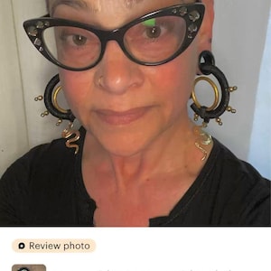 May include: Black ear weights with gold accents and a snake design. The ear weights are large and feature a unique, eye-catching design. The image also shows a person wearing black-rimmed glasses.