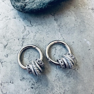 May include: Two silver hoop earrings with decorative beaded accents. Each earring features a thick, circular hoop with a central design of stacked, textured beads and smaller, black accent beads. The earrings are displayed on a textured gray surface.
