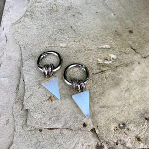 Puede incluir: Un par de pendientes plateados con una parte superior circular y un colgante triangular opalescente. Los pendientes se muestran sobre una superficie gris texturizada. Diseño único, adecuado para el uso diario.