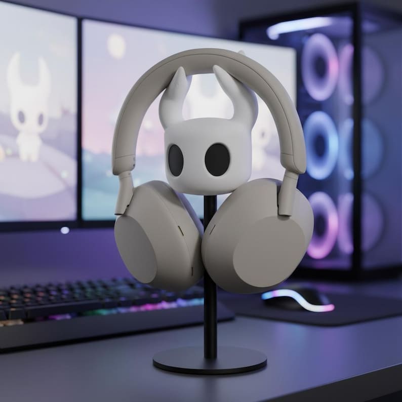 May include: A gray headset rests on a black headphone stand with a white character design. The stand has a circular base and a vertical support. The background includes a computer monitor and keyboard.