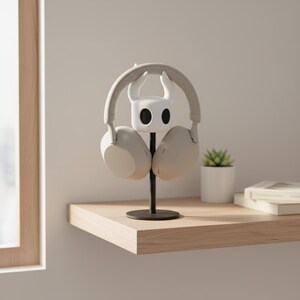May include: A white and black headphone stand shaped like a character from the video game Hollow Knight, holding a pair of light gray headphones. The stand is on a black metal pole, placed on a wooden shelf. A small potted plant and books are also on the shelf.