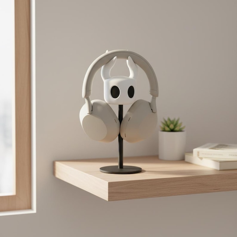 May include: A white and black headphone stand shaped like a character from the video game Hollow Knight, holding a pair of light gray headphones. The stand is on a black metal pole, placed on a wooden shelf. A small potted plant and books are also on the shelf.