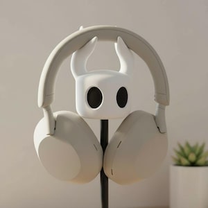 May include: A white headphone stand featuring a stylized character head with large black eye-like openings. The stand holds a pair of light gray over-ear headphones. The stand and headphones are set against a neutral background.