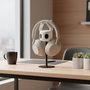 May include: A white headset rests on a black stand shaped like a character from the video game Hollow Knight. The stand is on a wooden desk, with a black mug and small potted plants in the background.