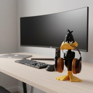 May include: A black Daffy Duck headphone stand with a yellow beak and feet, holding brown headphones. The stand is on a light wood desk, with a computer monitor, keyboard, and mouse in the background.