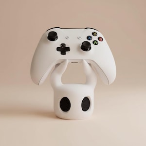 May include: A white Xbox controller rests on a white, stylized stand. The controller has black buttons and colored action buttons. The stand has a rounded shape with two large, black eye-like openings.