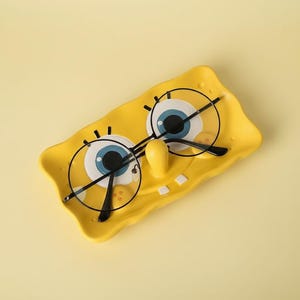 Op de afbeelding: Een gele keramische tray in de vorm van het gezicht van SpongeBob SquarePants, met grote blauwe en witte ogen en een gele neus. Een bril met zwart montuur ligt op de tray. De tray staat op een gele ondergrond.