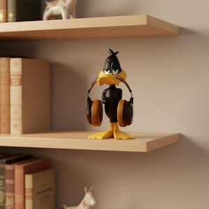 May include: A Daffy Duck figurine with a black body, yellow feet and beak, and brown headphones. The figurine is standing on a wooden shelf, with books and other decorative items in the background.