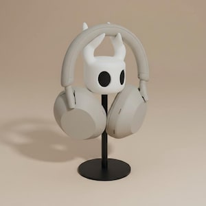 May include: A light gray headset rests on a black stand with a white character-shaped holder. The stand has a circular base and a vertical pole. The headset is a neutral color, and the holder has two black eye-like shapes.