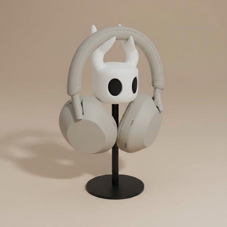 May include: A light gray headset rests on a black stand with a white character-shaped holder. The stand has a circular base and a vertical pole. The headset is a neutral color, and the holder has two black eye-like shapes.