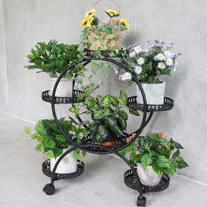 Peut inclure: Un présentoir à plantes en métal noir avec plusieurs étagères circulaires, chacune contenant des plantes et des fleurs en pot. Le présentoir est monté sur roues. Les plantes présentent un feuillage vert et des fleurs colorées.