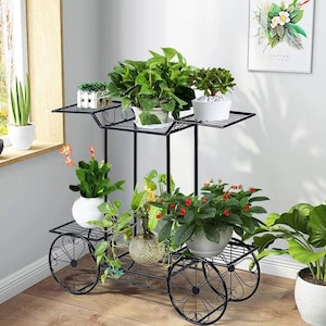 Puede incluir: Soporte para plantas de metal negro con tres niveles y detalles decorativos en forma de rueda. Exhibe varias plantas en macetas con follaje verde y flores rojas. El soporte se coloca cerca de una ventana, realzando la luz natural.
