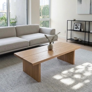 May include: A rectangular light brown wooden coffee table with a smooth surface and two thick slab legs. The table features a visible wood grain pattern and a horizontal seam running down the center of the top surface, supporting a small ceramic vase.