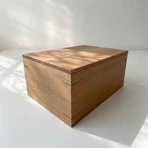 May include: A rectangular wooden box with a natural light brown wood grain finish. The box features a flat, removable lid that sits flush with the sides, showcasing visible joinery details at the corners of the structure.