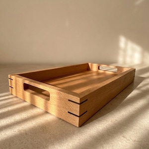 May include: A rectangular wooden tray with cut-out handles. The tray is made of light-coloured wood with visible grain and dark accents at the corners. The tray is empty and sits on a light-coloured surface.