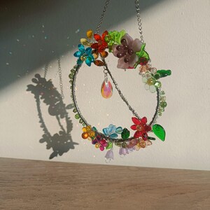 May include: A decorative floral wreath with a silver chain for hanging. The wreath is adorned with colorful, translucent flowers, green leaves, and a dangling iridescent crystal. The wreath is suspended in front of a white wall.