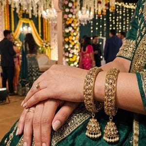Peut inclure: Gros plan sur des bracelets dorés avec des motifs ajourés complexes et des breloques en forme de cloche. Les bracelets ornent un poignet vêtu d'un vêtement vert et or brodé. L'image met en valeur les bijoux et la mode traditionnels.