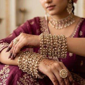 Puede incluir: Primer plano de un conjunto de joyas doradas. El conjunto incluye un collar, pendientes, anillos y varias pulseras. Las pulseras y el collar están adornados con pequeñas perlas y cristales transparentes. Los anillos son redondos con un centro de perlas. La modelo lleva un vestido granate.