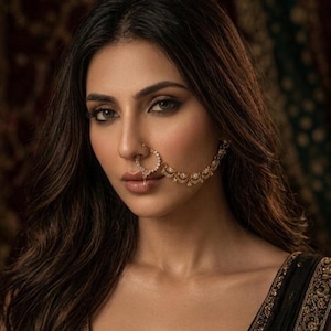 May include: A woman with long, dark brown hair and a gold nose ring and earring. The nose ring is a circular design with small, clear stones. The earring is a chain of gold with clear stones. The woman is wearing a black top with gold embroidery.