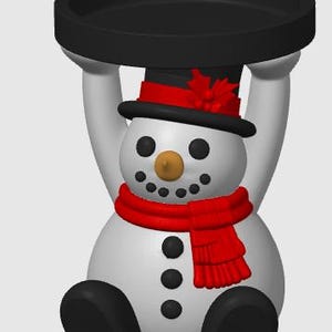 May include: A white snowman figurine with a black top hat featuring a red band and holly, a red scarf, and black buttons. The snowman is sitting with arms raised, supporting a circular black tray above its head. The snowman has a tan nose.