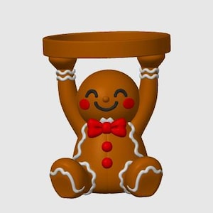 May include: A brown gingerbread person figurine holding a circular tray above its head. The figure features white icing-style trim, a red bow tie, two red buttons, and red cheeks. The gingerbread person has a smiling face with closed eyes.