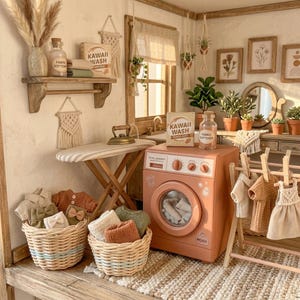 May include: A miniature laundry room scene with a peach-colored washing machine, ironing board, and baskets of clothes. The scene includes a wooden shelf with a bottle labeled "Fabric Softener" and a box of "Kawaii Wash". Decorative plants and framed art complete the cozy aesthetic.