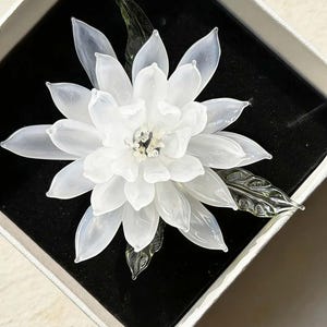 May include: A handcrafted glass flower brooch featuring translucent white petals arranged in multiple layers, a center with small clear glass beads, and two dark green glass leaves extending from the base, presented inside a square box.