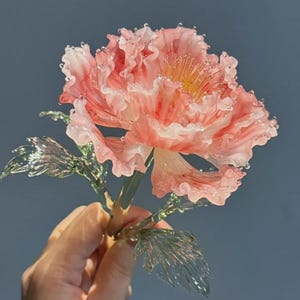 Op de afbeelding: Een handgemaakte glazen bloem met golvende, doorschijnende roze en witte bloemblaadjes en een geel hart. De bloem heeft delicate, heldergroene glazen bladeren aan een slanke steel, met gedetailleerde texturen en lichtreflecties.