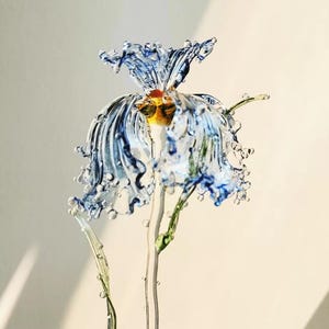 May include: A handcrafted glass flower sculpture featuring delicate, ruffled petals in shades of translucent blue and clear glass. The center displays warm yellow and orange accents, supported by a slender, clear glass stem with green leaves.